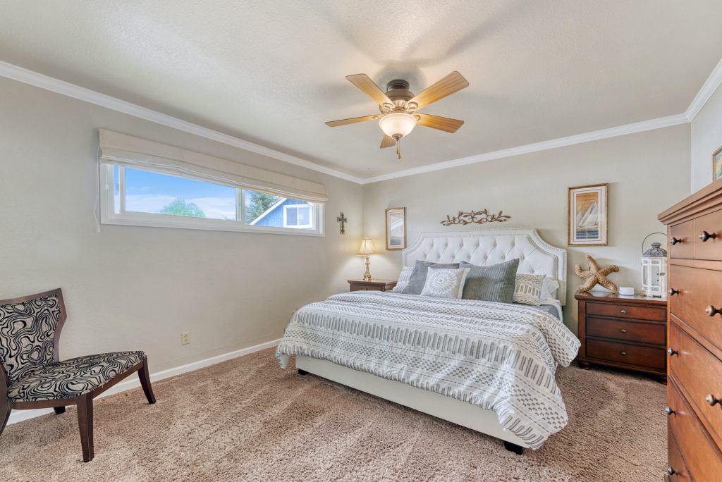 A bedroom with upscale furnishings, carpeting, ceiling fan and long narrow window